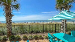 A beachside patio with a view of the ocean, featuring a striped umbrella, palm trees, and turquoise seating. - Sunset Palace Perfect Beach Getaway: Stunning Updates & Unbeatable Convenience! (Myrtle Beach)