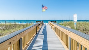Une promenade en bois menant à la plage, avec un drapeau américain et un panneau indiquant la présence d’un sauveteur.