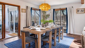 Un coin repas avec une table en bois dressée pour six personnes, entourée de chaises, agrémenté d’un luminaire suspendu et de grandes fenêtres avec des rideaux.