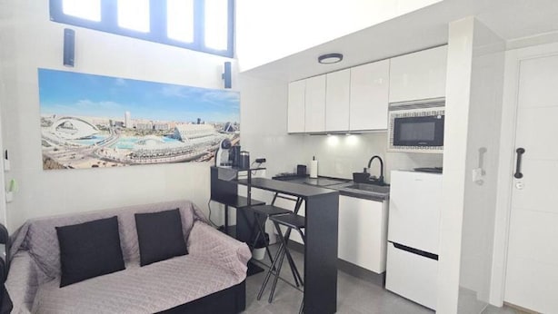 Un espace de vie compact comprenant un coin cuisine, un canapé avec des coussins, un coin repas et une grande fenêtre donnant sur le paysage urbain.