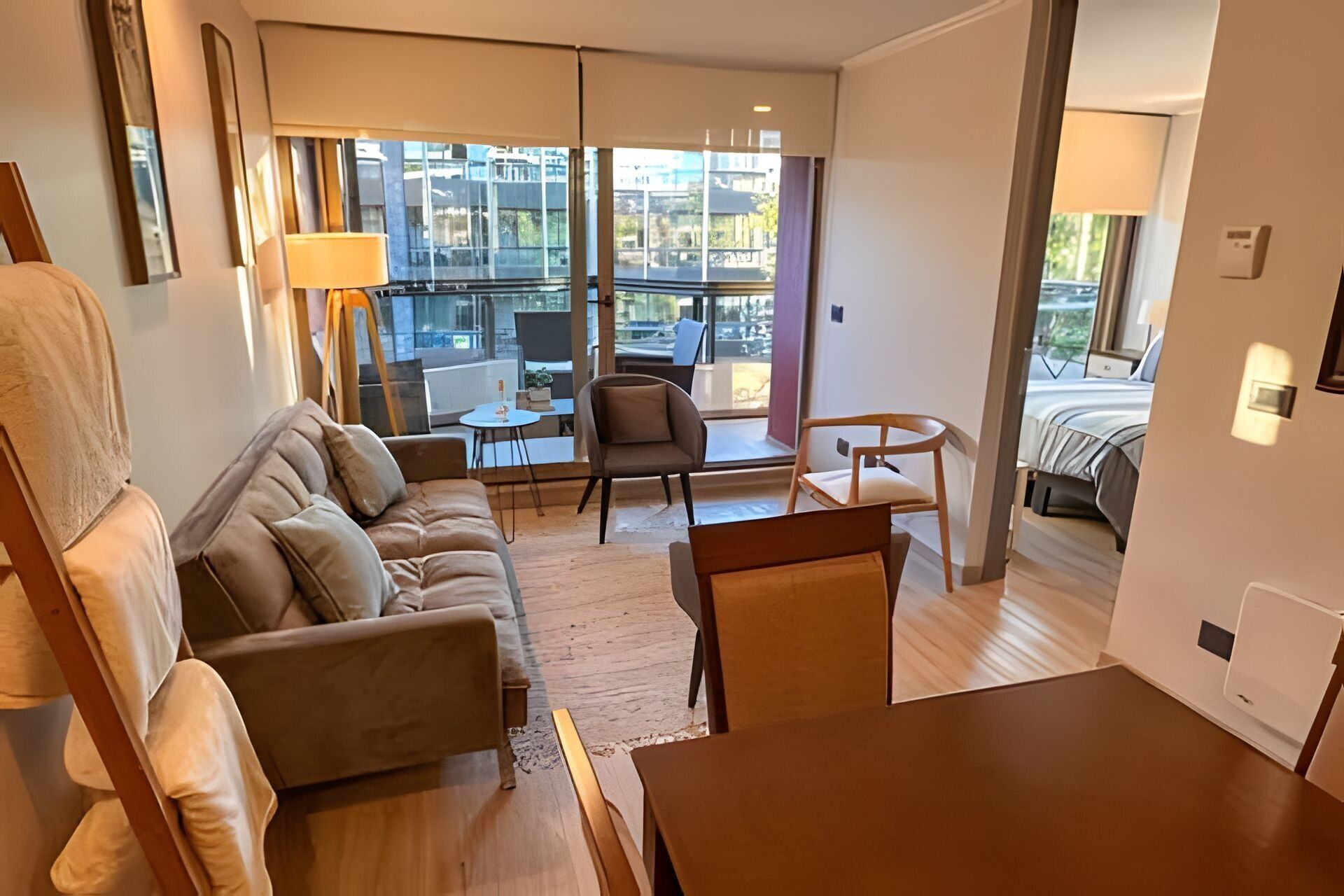 A modern hotel room with a sofa, chairs, a small table, and a view of the city through large windows.