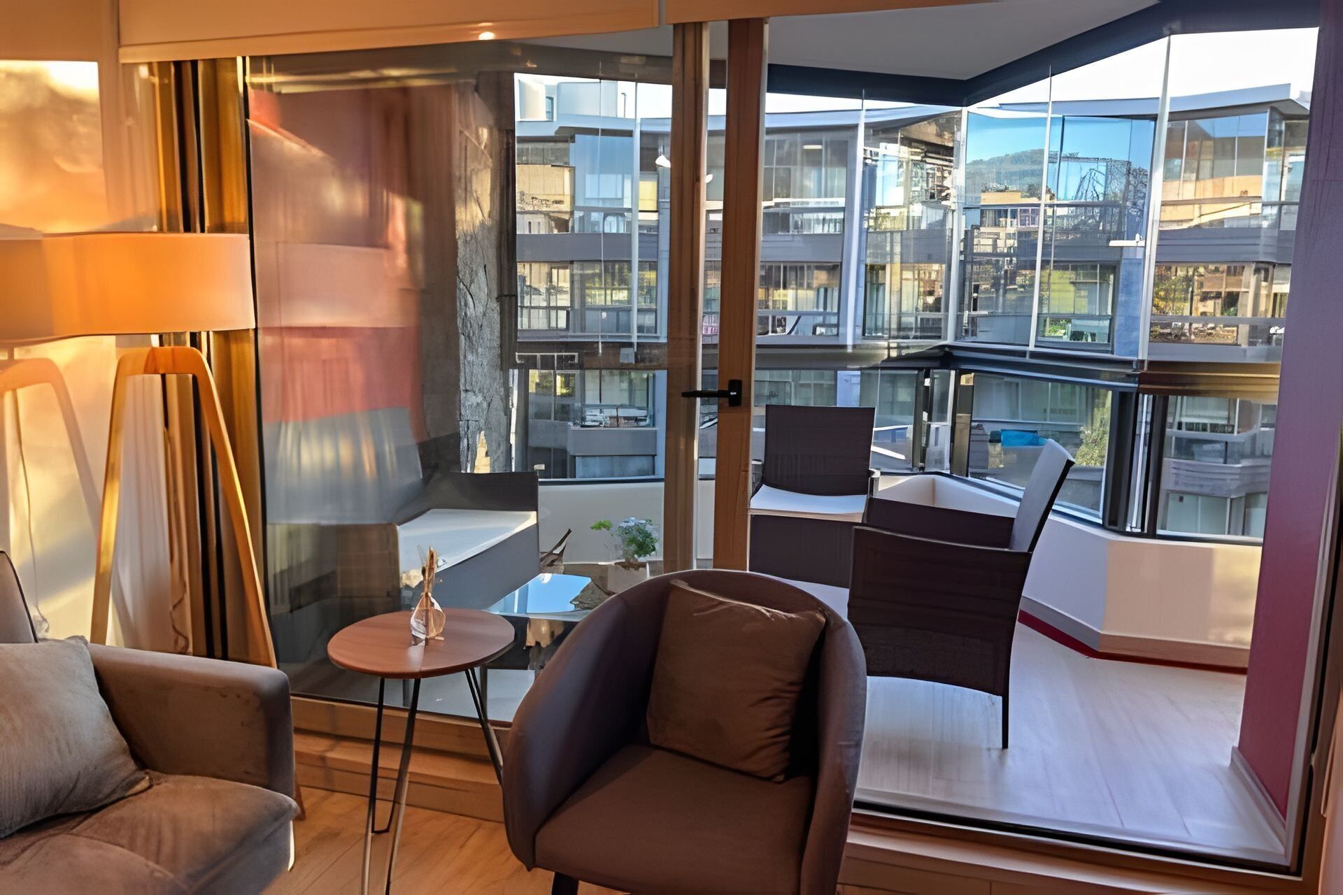 A modern living room with a sofa, armchair, and small table, a balcony with a view of a cityscape.