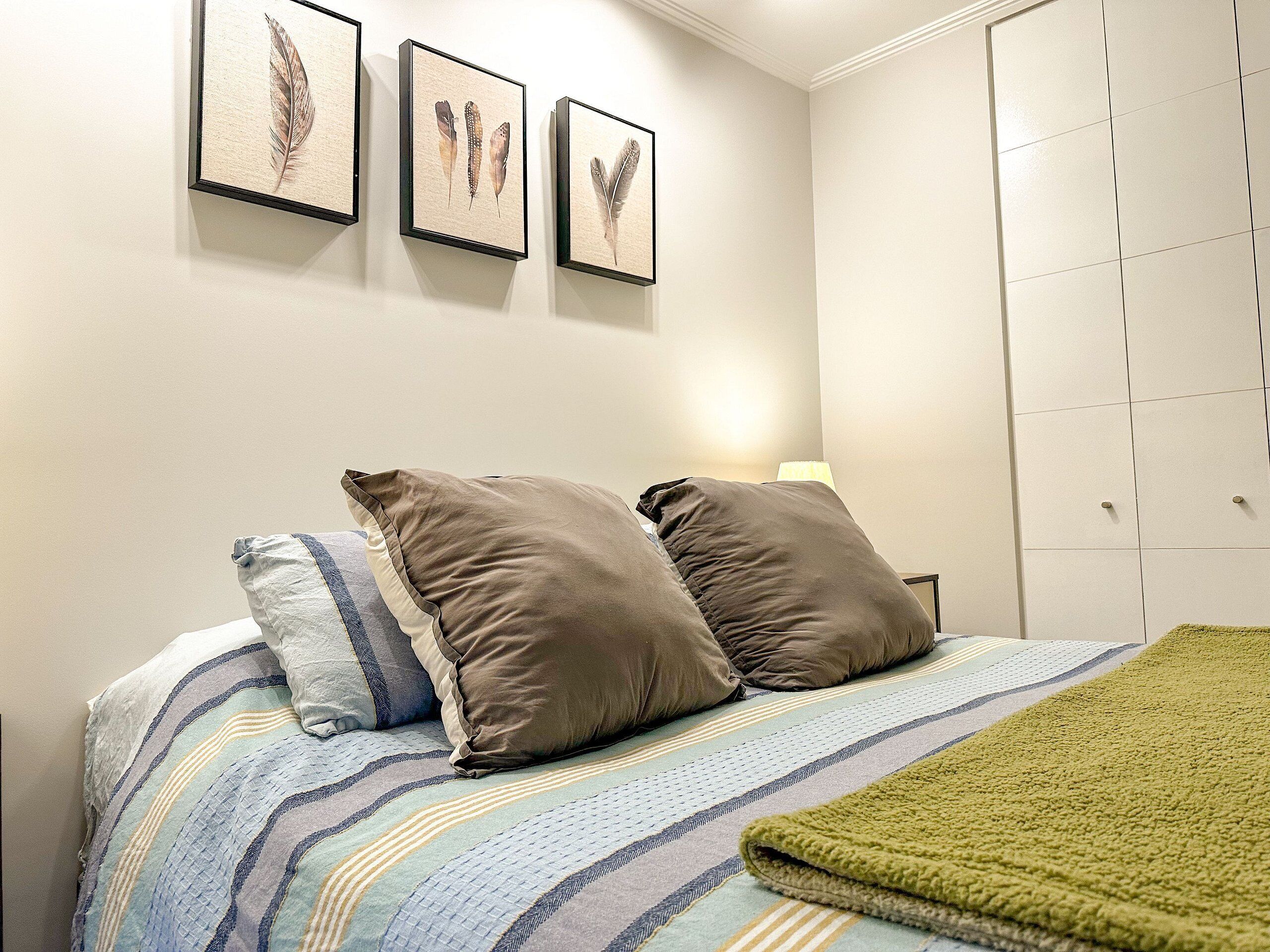 Una cama bien tendida con ropa de cama a rayas y dos almohadas, una manta verde y tres piezas de arte de plumas colgadas encima.