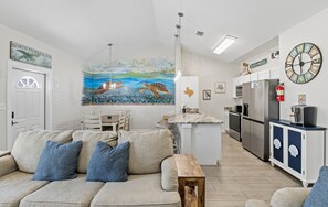 Una cocina moderna con una isla central, un comedor con un mural y una sala de estar con sofá y cojines.