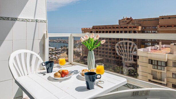 A balcony with a white table set for breakfast, including croissants, fruit, and orange juice, with a view of the city and sea.