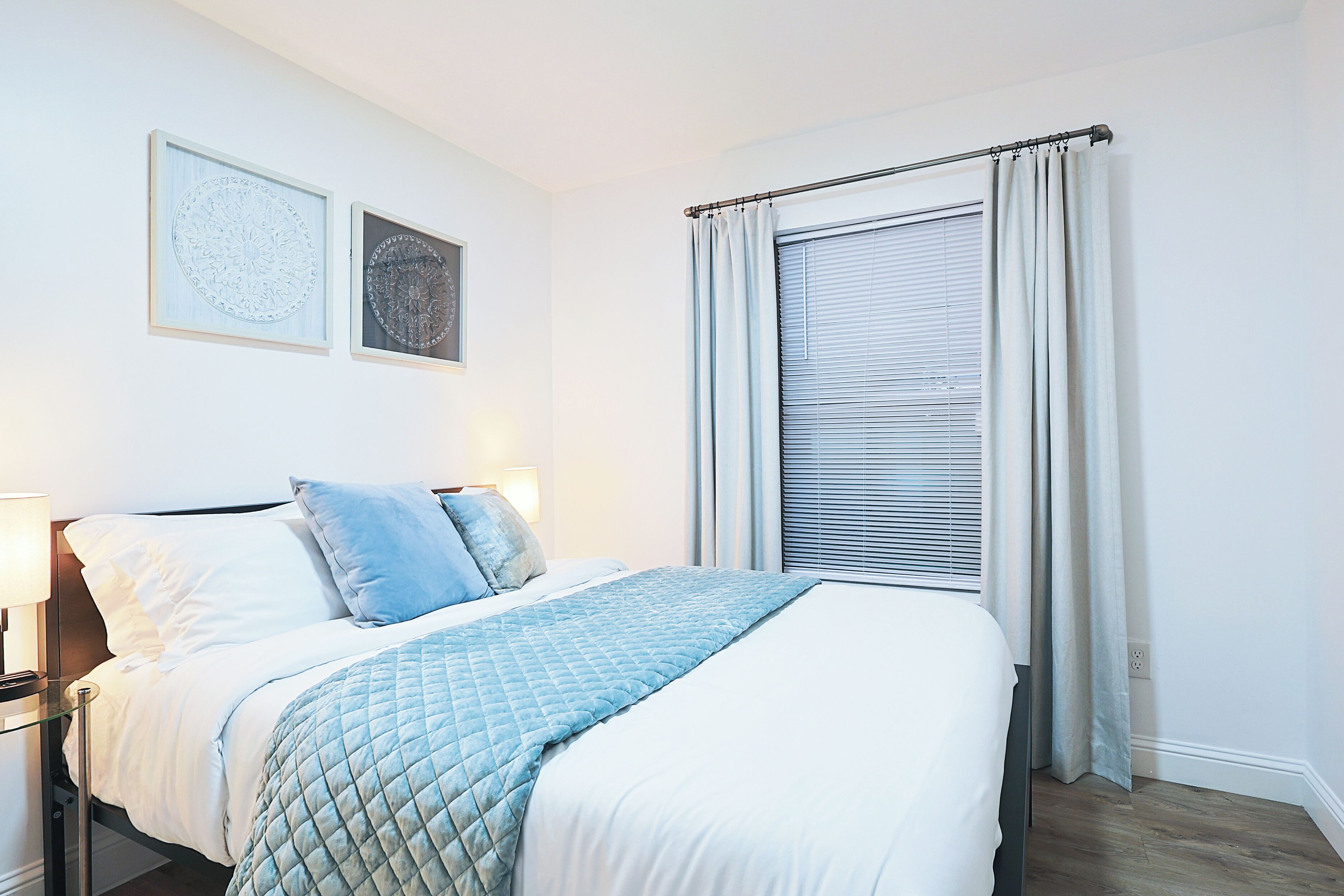 Een net opgemaakt bed met een gewatteerde deken, een bedlampje en twee ingelijste kunstwerken aan de muur.