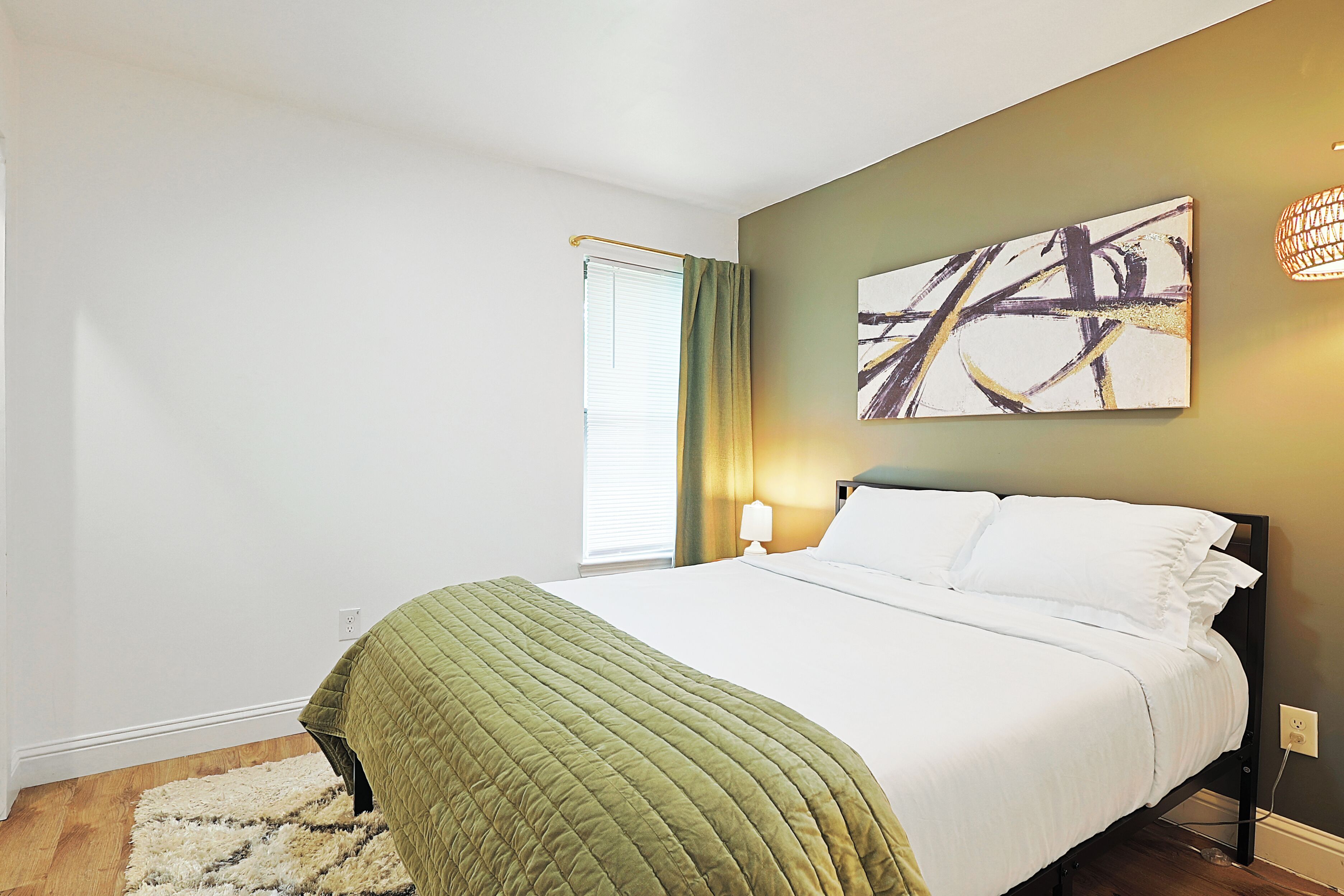 A neatly made bed with a green quilt, a wall-mounted abstract painting, and a bedside lamp.