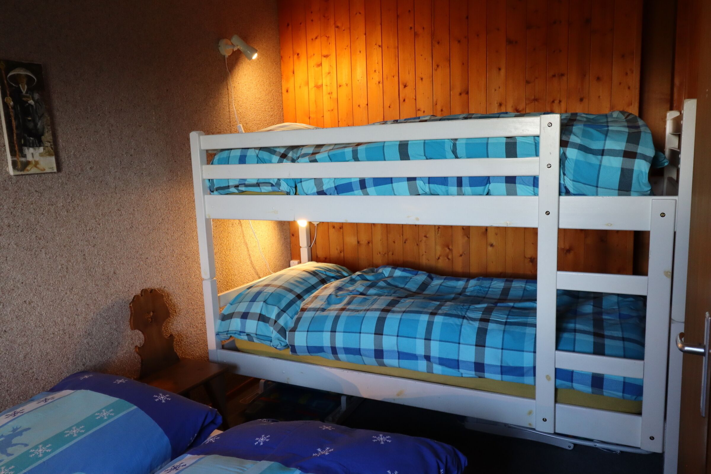 Een stapelbed met blauw-witte beddengoed, houten wanden en een klein nachtkastje met een lamp.