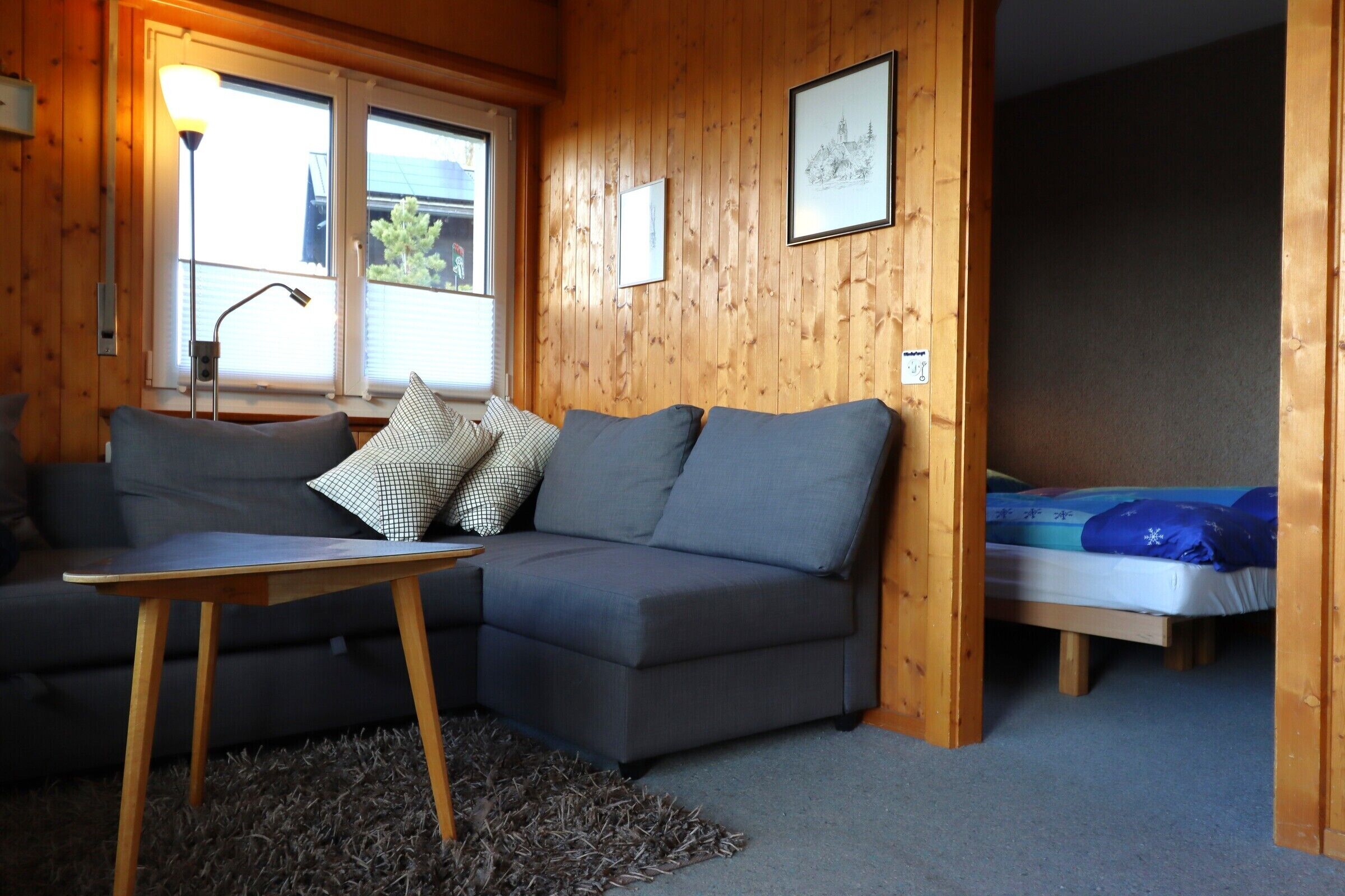 Ein gemütliches Zimmer mit einem grauen Sofa, einem kleinen Holztisch und einem Bett mit blauer Decke.