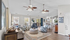 A spacious living room with a large round ottoman, two ceiling fans, and a dining area with a chandelier. - Charming 3BR, 2BA Canal-Front Getaway with Kayaks in Oak Island, NC! (Oak Island)