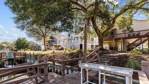 A wooden deck with a table and benches, surrounded by trees and a small water feature. - Charming 3BR, 2BA Canal-Front Getaway with Kayaks in Oak Island, NC! (Oak Island)