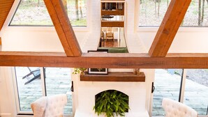 Quarto com lareira, dois poltronas brancas, teto de madeira e grandes janelas com vista para uma floresta.