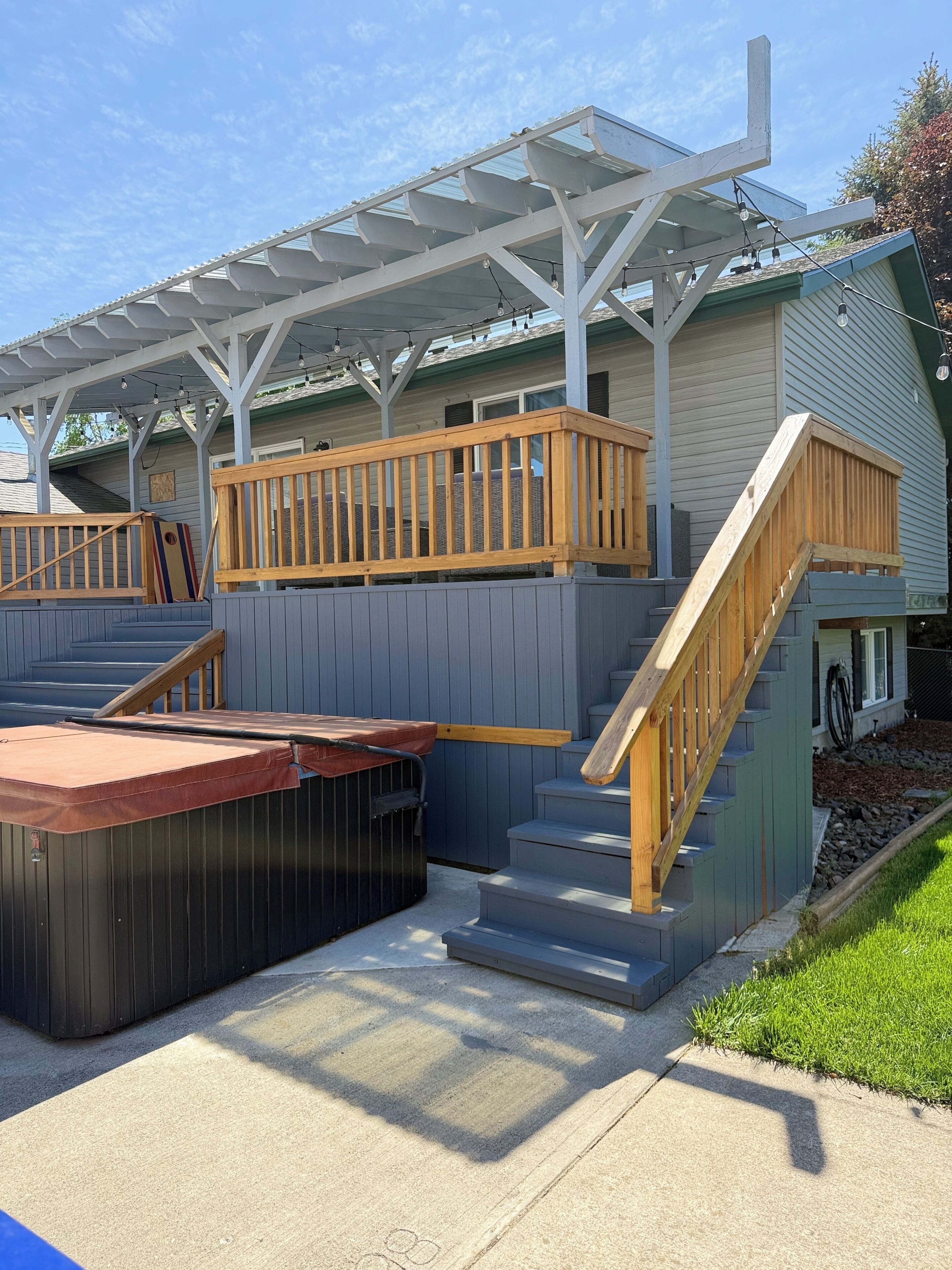 A covered outdoor area with a hot tub, wooden deck, and stairs leading to a house.