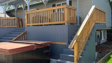 A covered outdoor area with a hot tub, wooden deck, and stairs leading to a house.