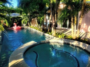 A swimming pool with a water fountain, surrounded by palm trees and a tiled deck.