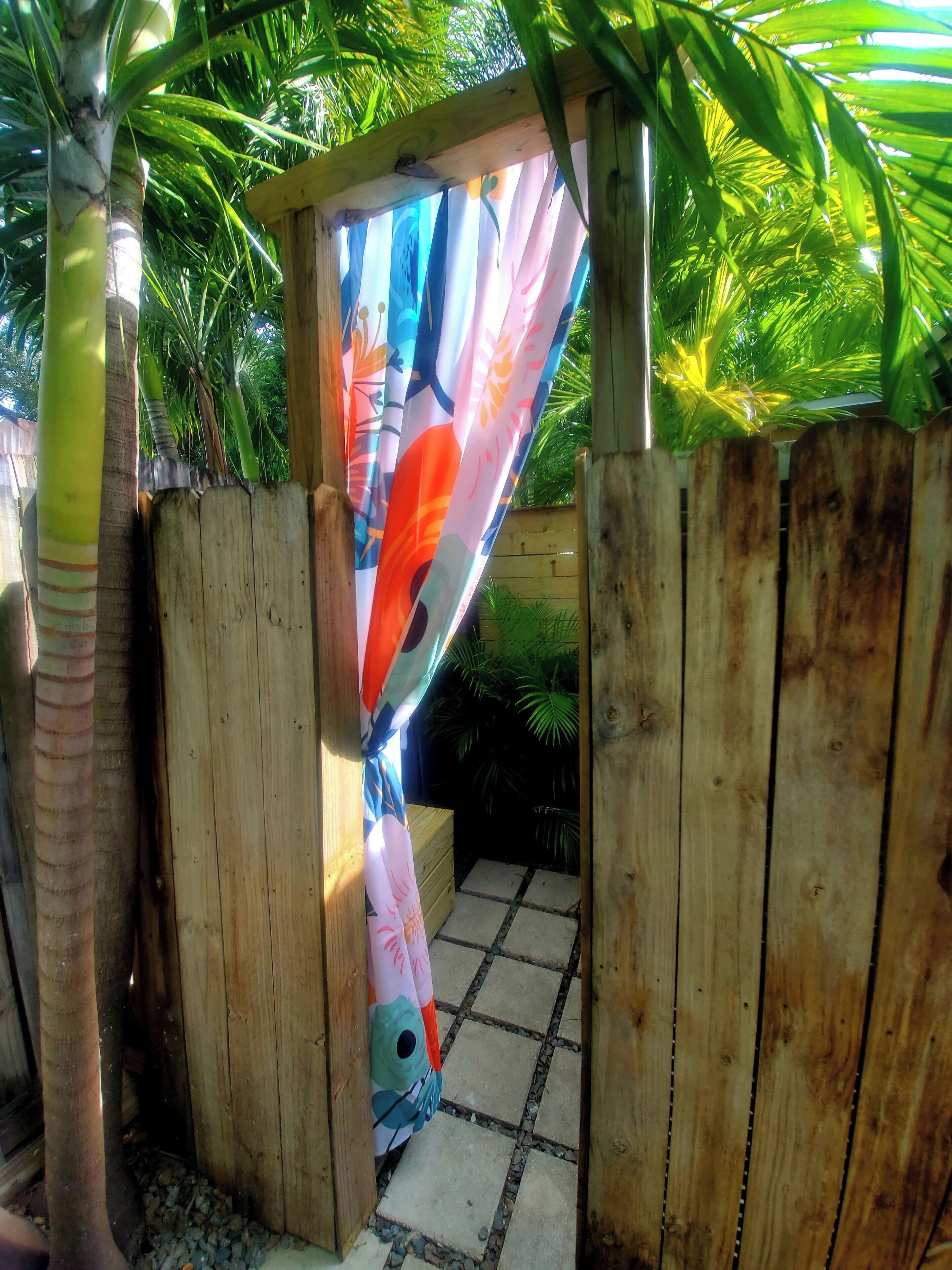 A wooden gate with a colorful fabric curtain, a palm tree, and a stone pathway leading into a shaded area.