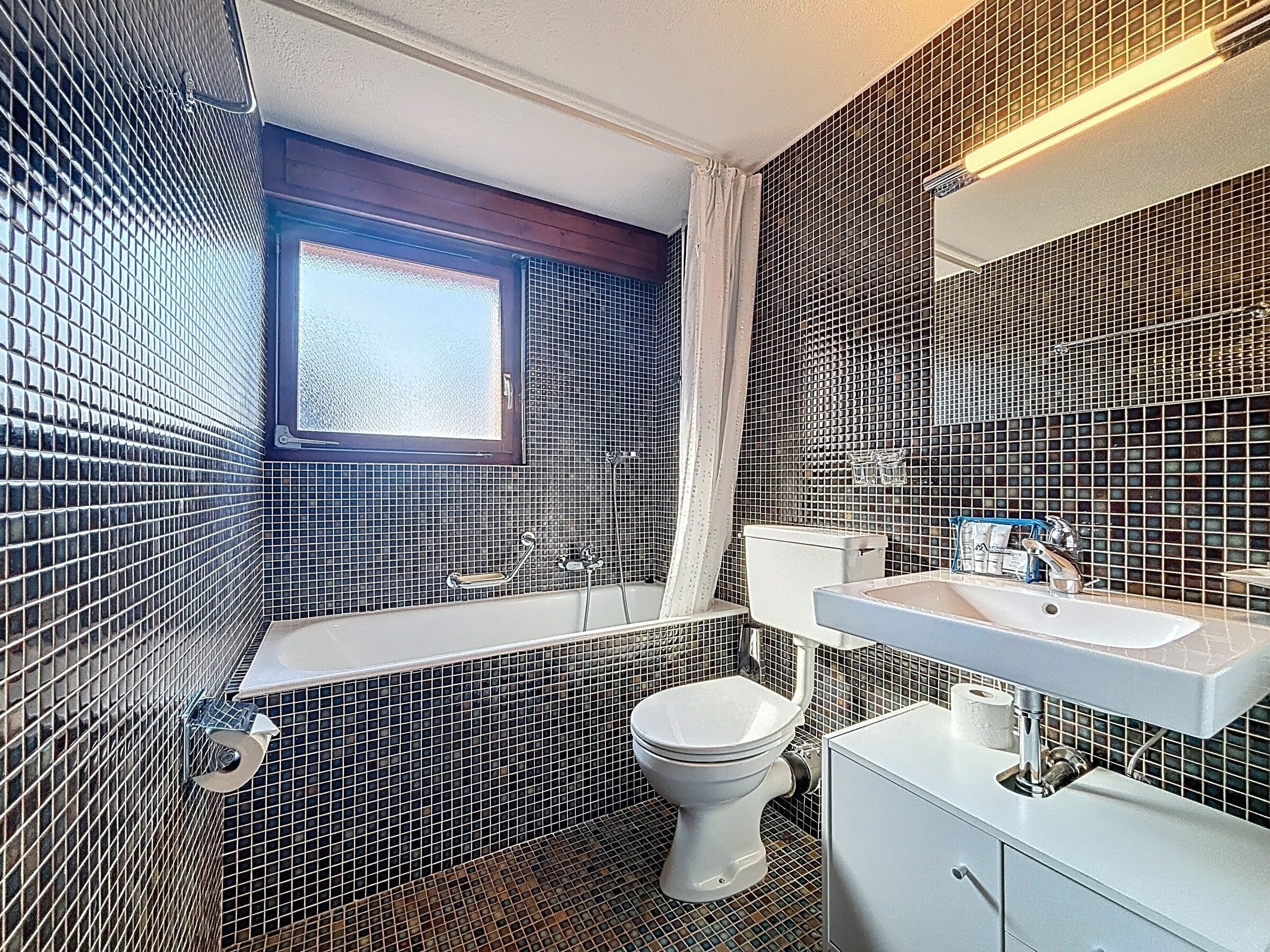 A bathroom with a bathtub, toilet, and sink, featuring black and white mosaic tile walls and a window.