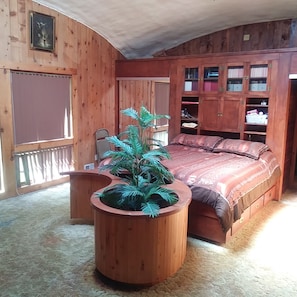 Un dormitorio con cabecera de madera, cama con colcha estampada, una planta en maceta y estanterías empotradas de madera con cajas de almacenamiento.