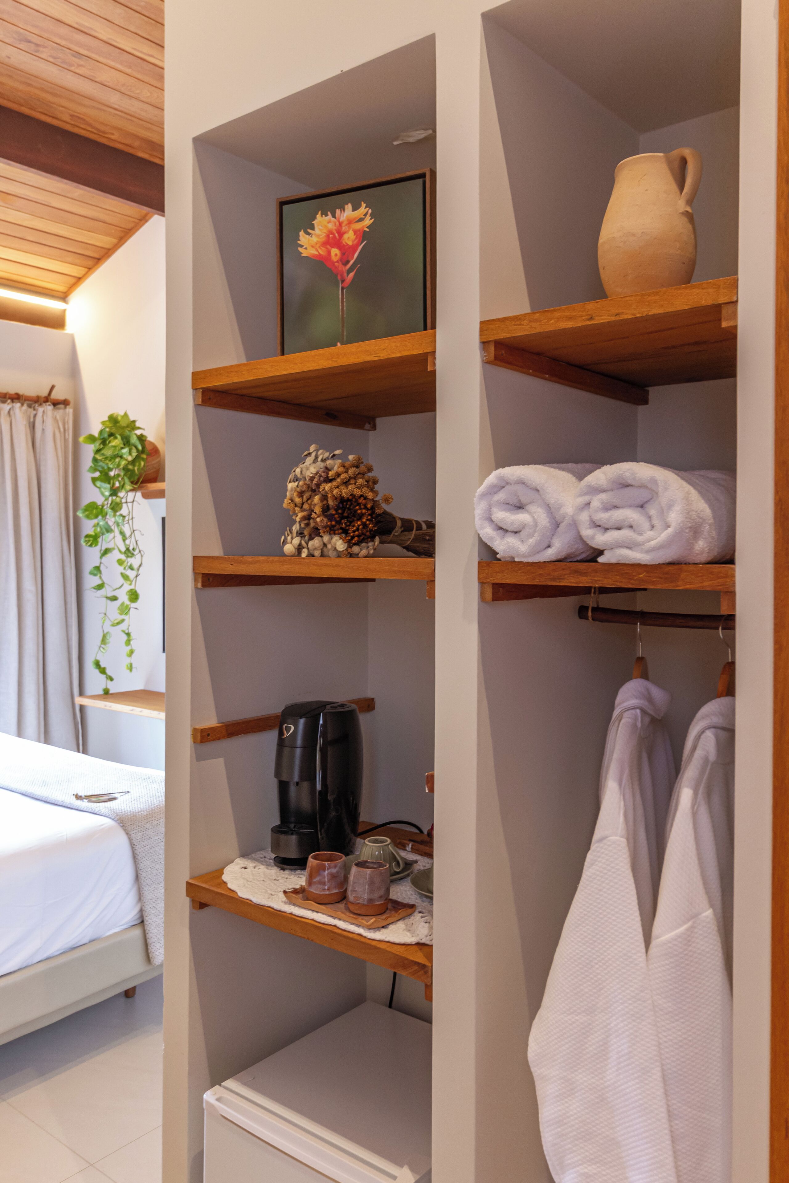 Una habitación ordenada con una cama, un estante con toallas, una cafetera y una planta en maceta.