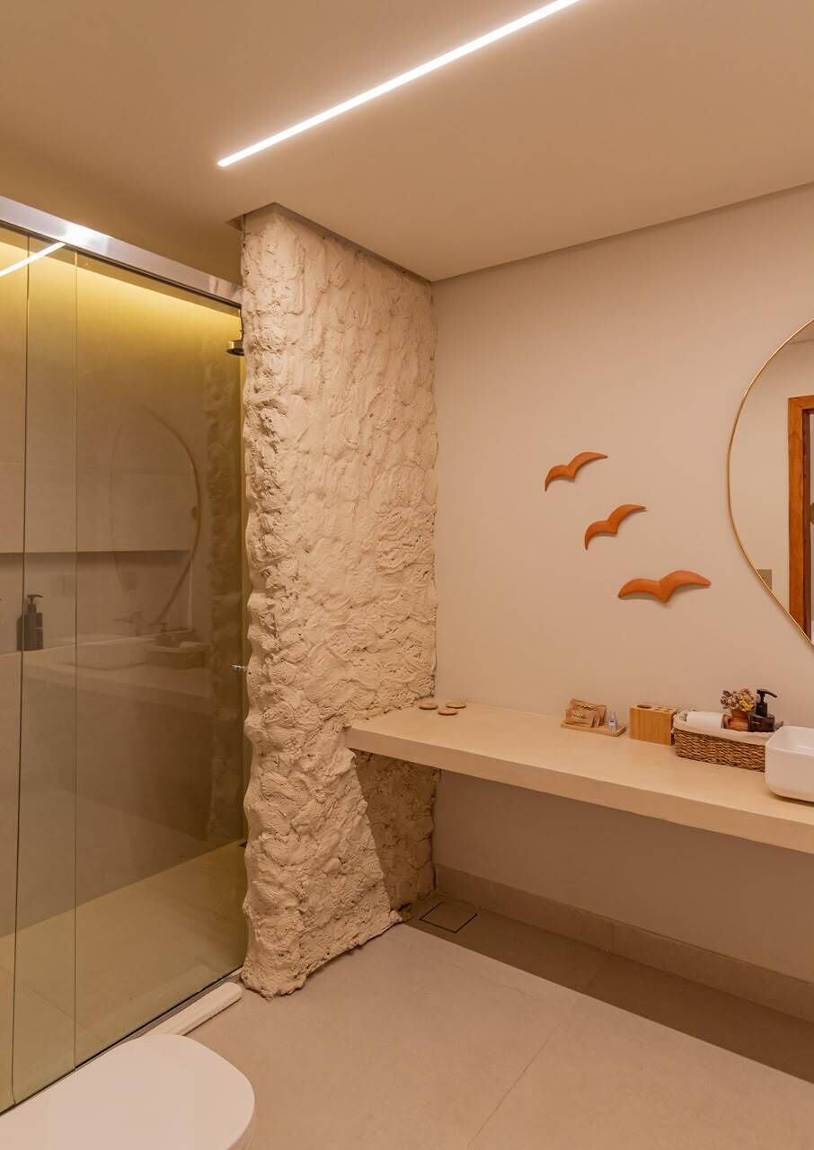 Un baño con cabina de ducha de vidrio, pared decorativa de piedra, espejo con marco circular y repisa con artículos de aseo.