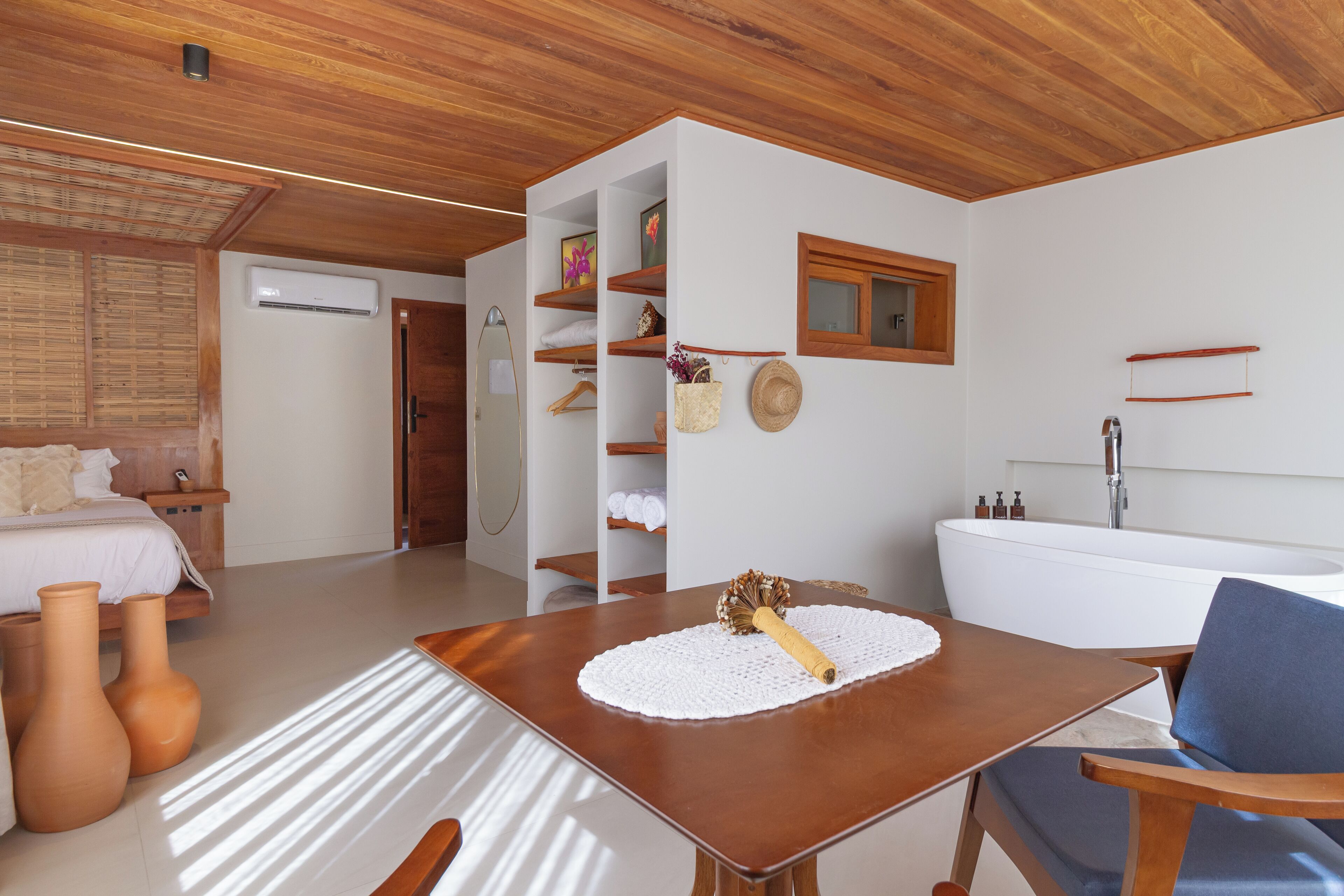Quarto moderno com teto de madeira, ar condicionado branco, uma banheira grande e branca, e uma mesa de madeira com um jogo americano trançado.