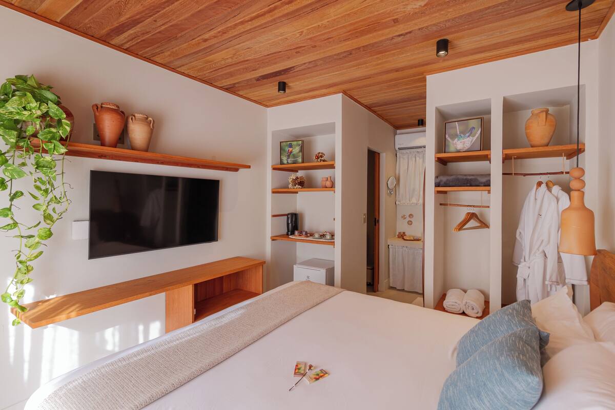 A modern hotel room with a wooden ceiling, a flat-screen TV mounted on the wall, a bed with white linens, a bedside table, and a plant.