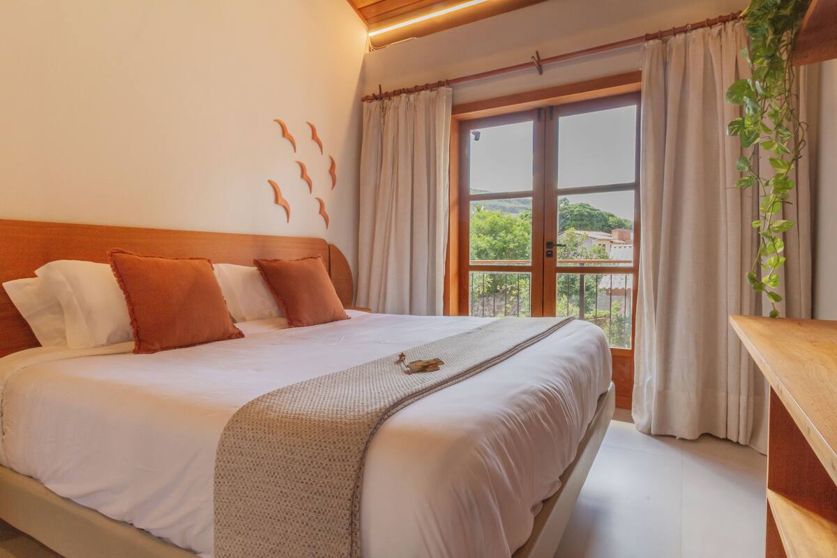 A bedroom with a large bed, a wooden headboard, a window with curtains, and a plant on a wooden table.