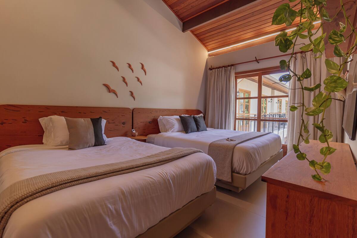 A hotel room with two beds, wooden headboards, a wooden desk, and a view of the outside.