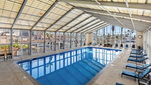 Una piscina interna con pareti in vetro trasparente, acqua azzurra e soffitto illuminato dal sole.