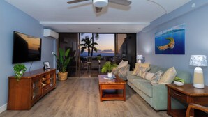 Ein Wohnzimmer mit Deckenventilator, einem Flachbildfernseher, einem grünen Sofa, einem Holztisch und Blick auf den Ozean durch große Glasfenster.