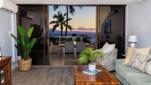 A living room with a view of palm trees and the ocean, a wooden coffee table, a sofa, and a dining area with chairs. - Oceanview Kahana Villa - Walk to Beach & Dining (Lahaina)
