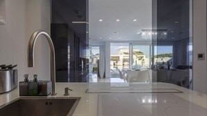 A modern kitchen with a stainless steel sink, a large curved faucet, and a glass partition leading to a dining area with a white table and chairs. - Luxury Sea View Villa with Pool in Santa Maria del Focallo – Villa Barbara, Sici (Ispica)