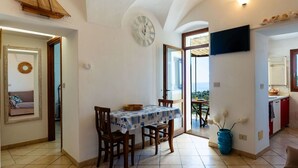 Un coin repas avec une table et des chaises, une horloge au mur et une vue sur la mer par une porte ouverte.