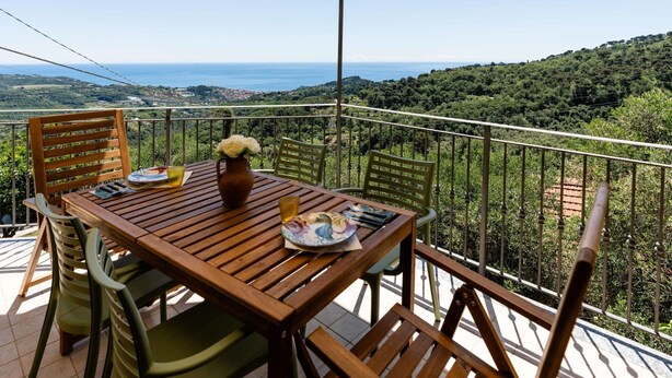 Une table en bois, entourée de chaises, sur un balcon offrant une vue panoramique sur des collines et la mer.