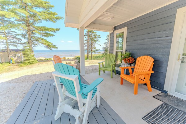A covered porch with Adirondack chairs and a table, overlooking a beach and ocean.