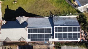 An aerial view of a building with a large solar panel array on the roof, a garage, and a fenced yard. - Pousada DonaBella (Campos do Jordão)