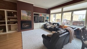 Een woonkamer met een open haard, een zithoek, een boekenkast en grote ramen.