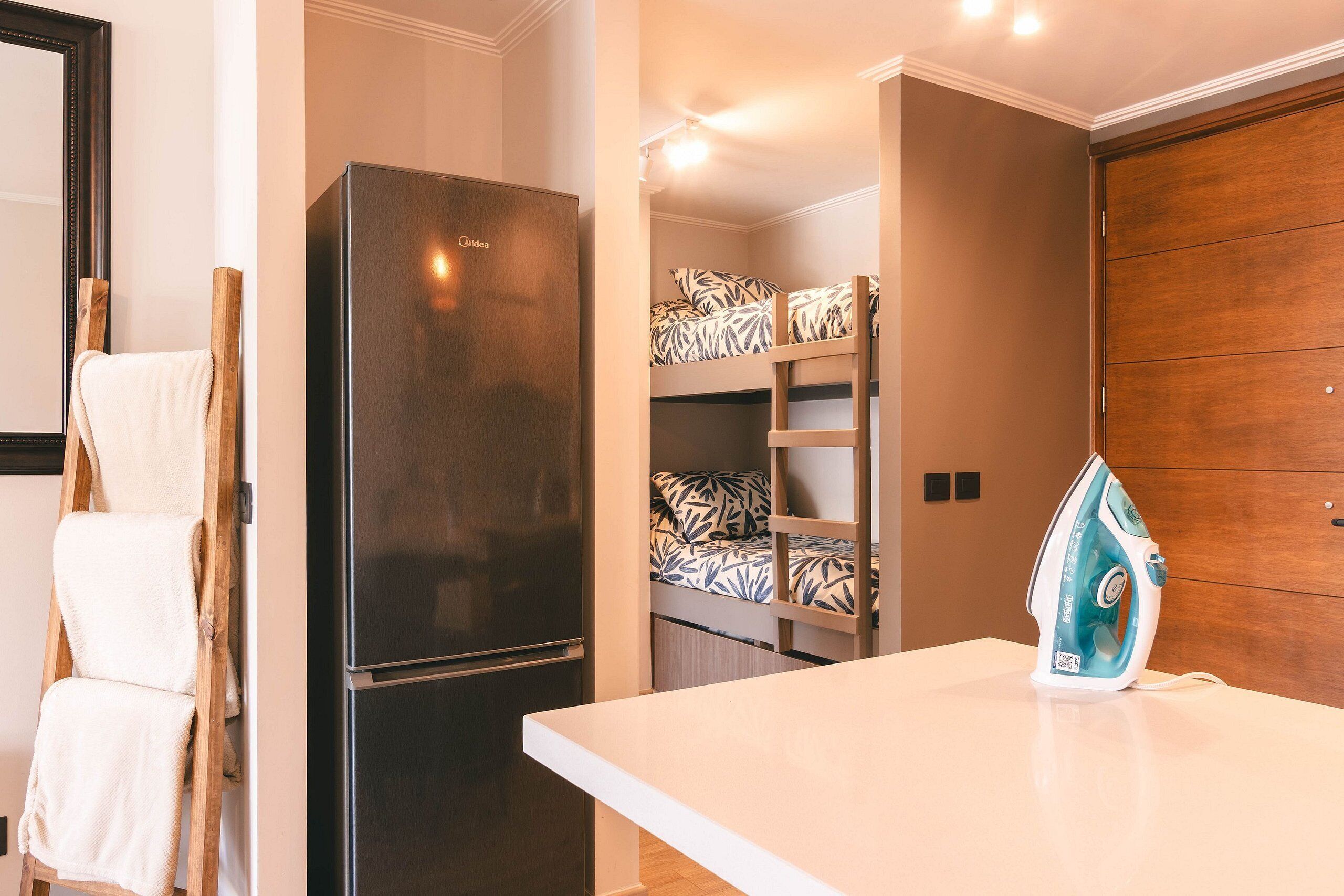 Una cocina compacta con refrigerador incorporado, una tabla de planchar de madera y una encimera blanca.