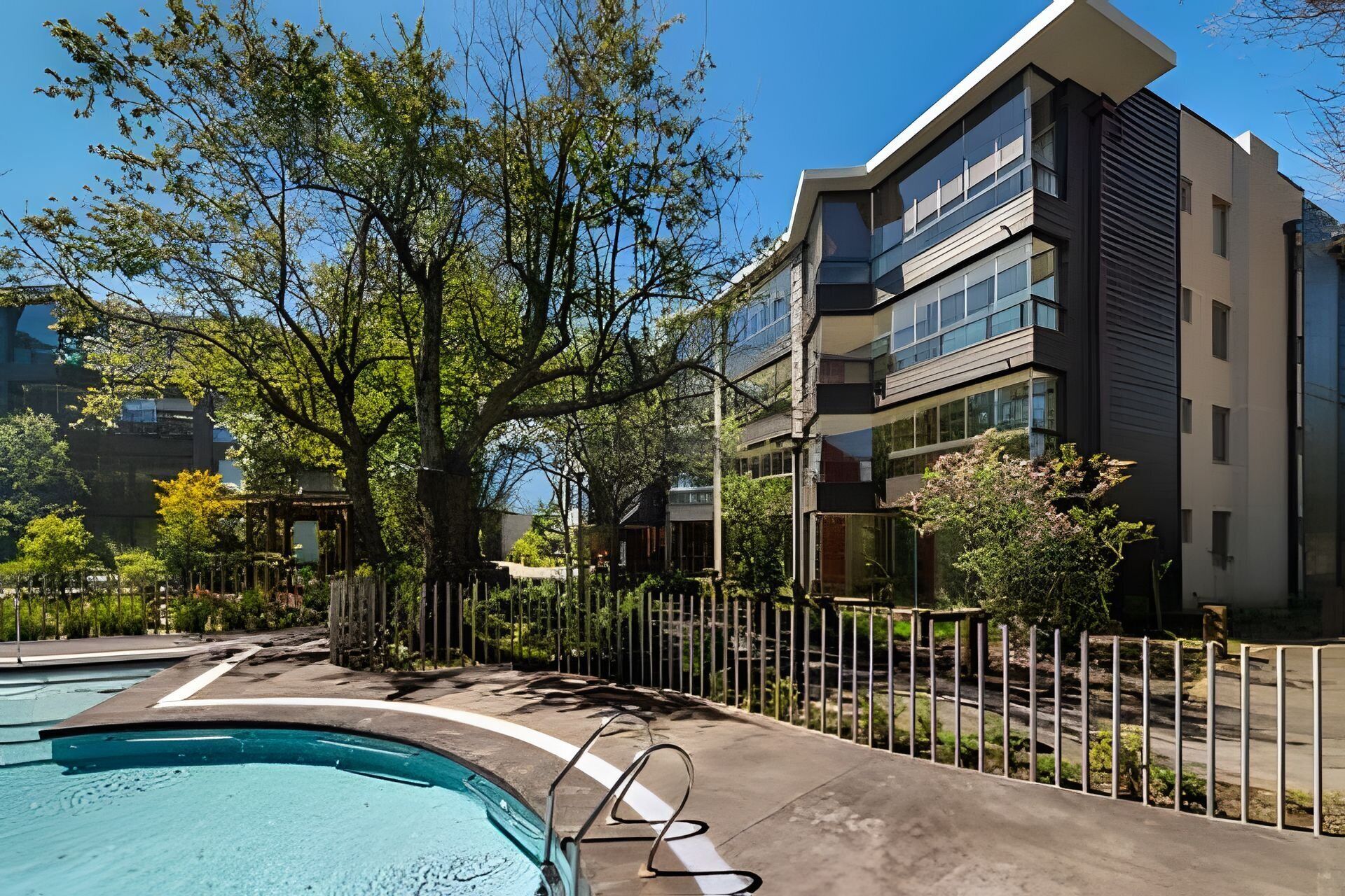 A modern building with a swimming pool, surrounded by trees and a metal fence.