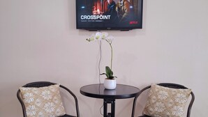 Una habitación con un televisor mostrando 'CROSSPOINT' en la pantalla, dos sillones con cojines estampados, una mesita con una orquídea en maceta y un televisor montado en la pared.