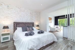 Une chambre moderne avec un grand lit, deux tables de chevet, une commode, une chaise et une vue sur la verdure.