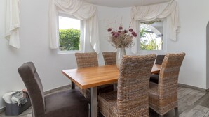 Un comedor con una mesa de madera y sillas de mimbre, un jarrón con flores secas y cortinas blancas.