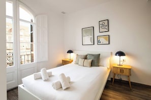 Una cama bien hecha con sábanas blancas y toallas enrolladas, dos lámparas de noche y dos cuadros en la pared.