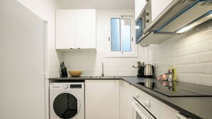 A compact kitchen with white cabinets, a built-in washing machine, a stove, and a window with blue-tinted glass.