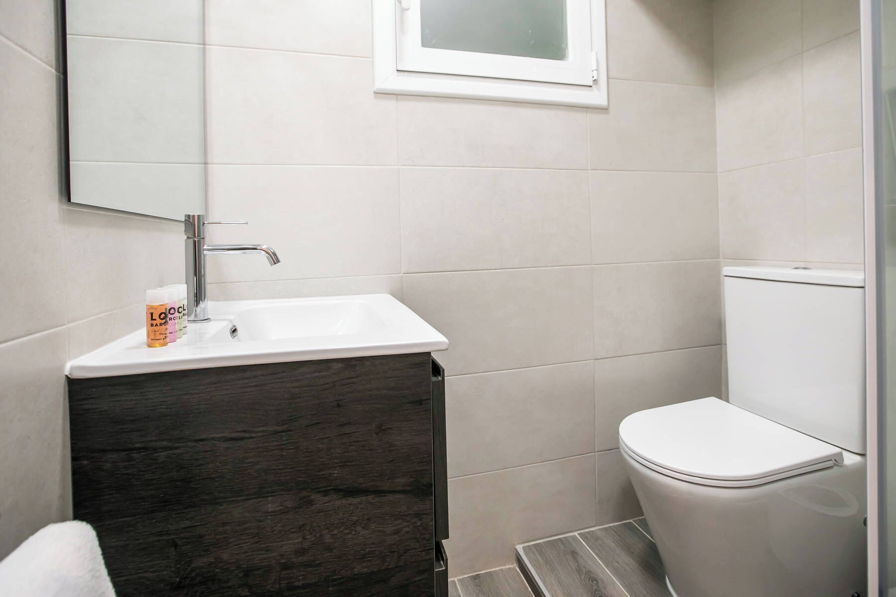 A bathroom with a white sink, dark wood vanity, white toilet, and a small window above the sink.