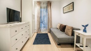 Ein gemütlicher, kleiner Wohnraum mit Bett, Fernseher auf einer Kommode, Schreibtisch mit Stuhl und einem gerahmten Bild an der Wand.
