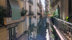 Une rue étroite en milieu urbain, bordée de bâtiments des deux côtés, avec des balcons ornés de plantes et une banderole publicitaire annonçant un événement.