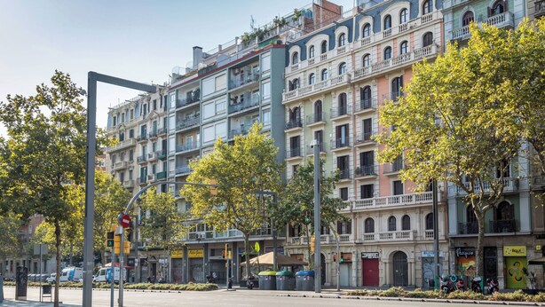 A multi-story building with balconies, a street with trees, and a clear sky.