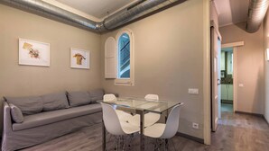 A modern living room with a glass-top dining table, white chairs, a grey sofa, and two framed artworks on the wall.