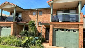 A two-story house with a brick exterior, a balcony on the upper floor, and a green garage door. - Shellharbour Beach Daze (Shellharbour)